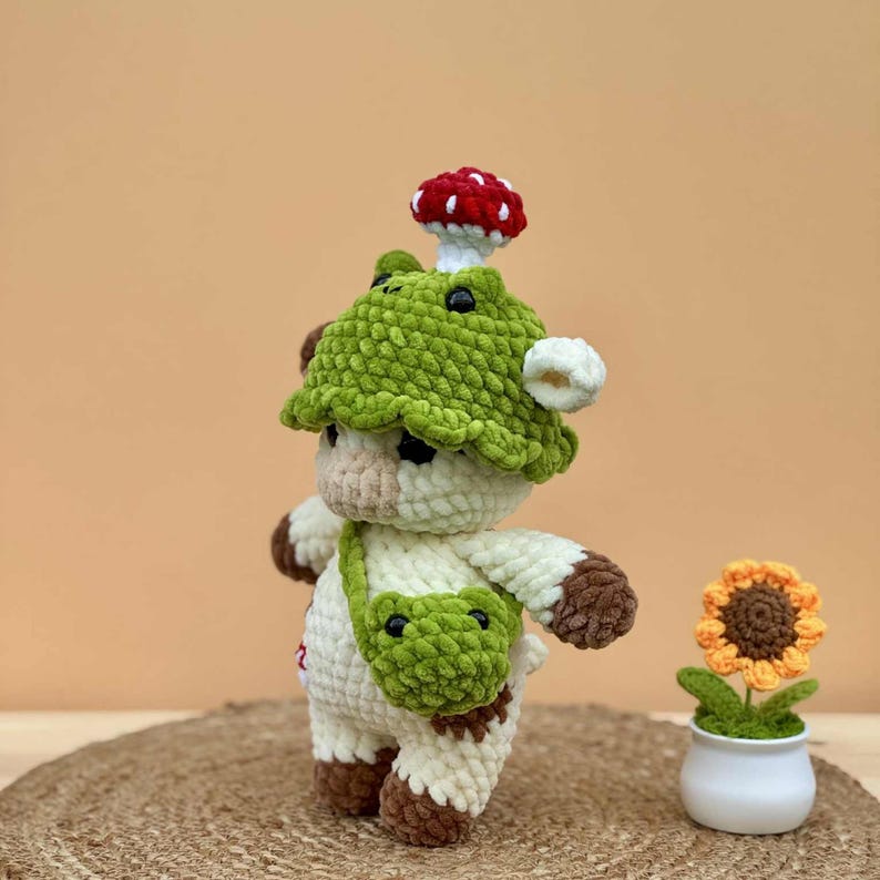 May include: A handmade crochet cow figurine with a green mushroom hat and a small green frog-shaped bag. The cow is white with brown accents and a red and white mushroom on top. A crochet sunflower in a white pot is also present.