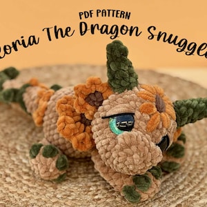 Può includere: Un giocattolo a forma di drago di peluche nei toni del beige, arancione e verde, con il testo "PDF PATTERN Floria The Dragon Snuggler". Il drago ha grandi occhi espressivi e un aspetto morbido e strutturato.