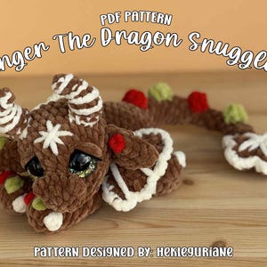 May include: A brown crocheted dragon plush with white icing-like embroidery, red and green accents, and large dark eyes. The text reads PDF PATTERN Ginger The Dragon Snuggler and PATTERN DESIGNED BY: HEKLEGURIANE. The dragon has horns and a long tail.