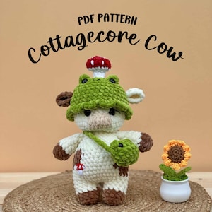 May include: A crocheted cow doll with a green frog hat and a mushroom on top. The cow has brown spots and a green satchel. A crocheted sunflower in a white pot is to the right. The text "PDF PATTERN Cottagecore Cow" is at the top.