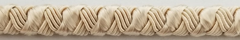 May include: A close-up of a cream-colored braided trim. The trim is made of a soft, woven fabric and has a delicate, intricate design.