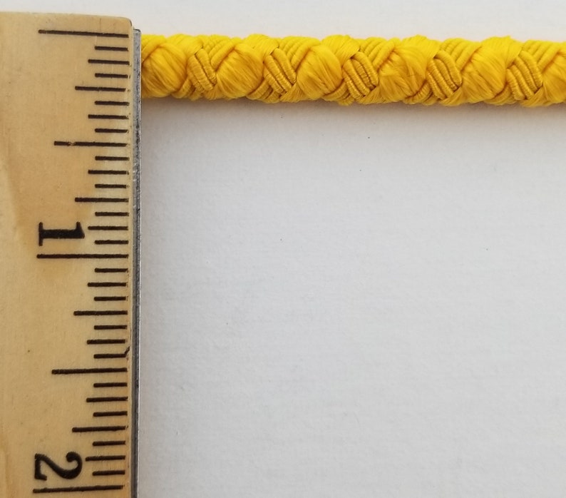 May include: A close-up of a bright yellow braided cord, measured against a ruler. The cord is approximately 1 inch wide.