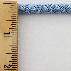 May include: A close-up of a light blue braided trim measured against a ruler. The trim is approximately 1 inch wide.