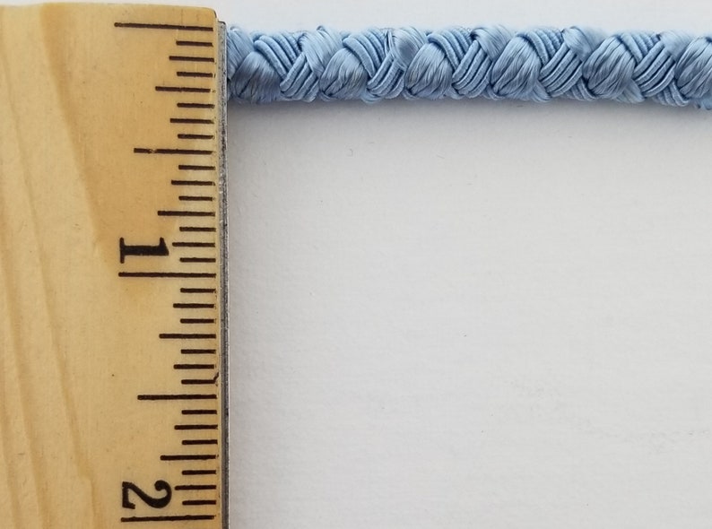 May include: A close-up of a light blue braided trim measured against a ruler. The trim is approximately 1 inch wide.