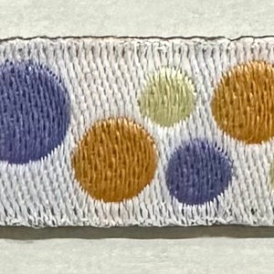 Puede incluir: Una cinta blanca con un patrón repetido de círculos bordados en tonos de azul, amarillo y naranja. La cinta tiene una apariencia texturizada y es adecuada para manualidades o adornos.