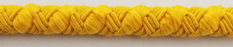 May include: A close-up of a yellow braided trim. The trim is made of a soft, woven fabric and has a slightly textured surface.