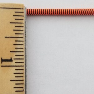 May include: A close-up of a black and orange striped elastic cord measured against a wooden ruler. The ruler shows 1 inch.
