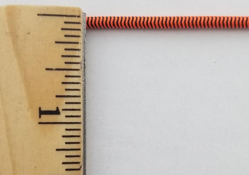 May include: A close-up of a black and orange striped elastic cord measured against a wooden ruler. The ruler shows 1 inch.