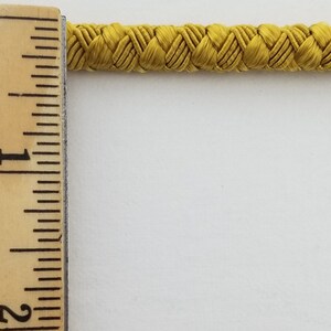 May include: A close-up of a yellow braided cord, measured against a ruler. The cord is approximately 1 inch wide.