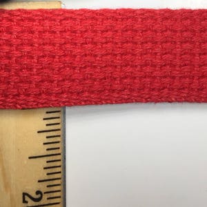 May include: Close-up of a red woven cotton webbing strap. The strap is measured with a ruler showing the 2-inch mark.