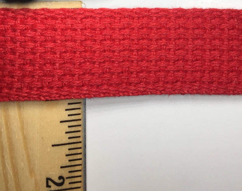 May include: Close-up of a red woven cotton webbing strap. The strap is measured with a ruler showing the 2-inch mark.