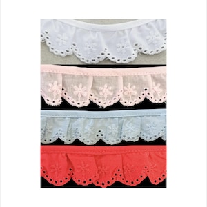 May include: Four strips of ruffled eyelet trim arranged vertically. The colors from top to bottom are white, pale pink, light blue, and coral red. Each strip features a scalloped edge with embroidered floral patterns and small circular cutouts.