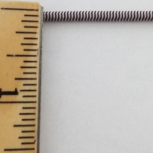 May include: A close-up of a white and black striped elastic cord measured against a wooden ruler. The ruler shows 1 inch.