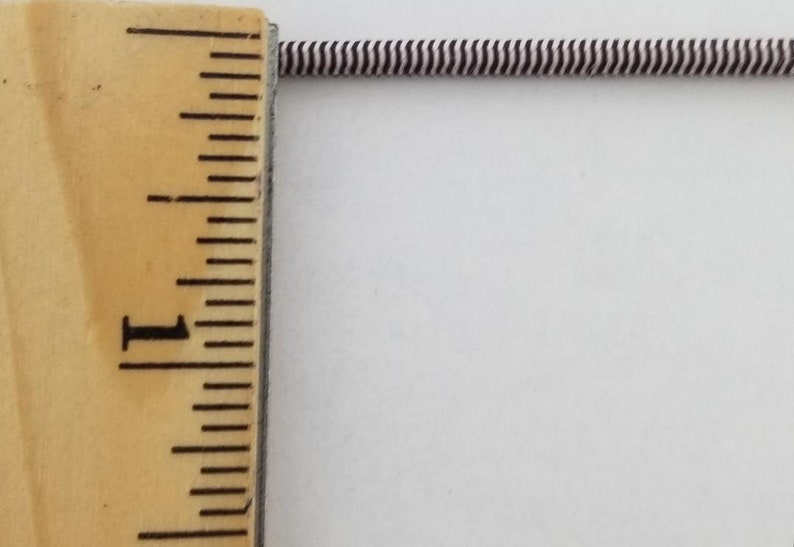 May include: A close-up of a white and black striped elastic cord measured against a wooden ruler. The ruler shows 1 inch.