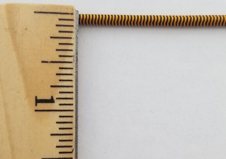 May include: A close-up of a black and yellow striped elastic cord, measured against a wooden ruler with inch markings. The cord is approximately 1 inch long.