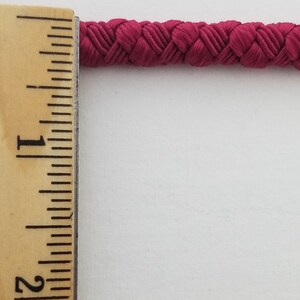 May include: A close-up of a 1-inch wide, crimson red braided cord. The cord is measured against a wooden ruler.