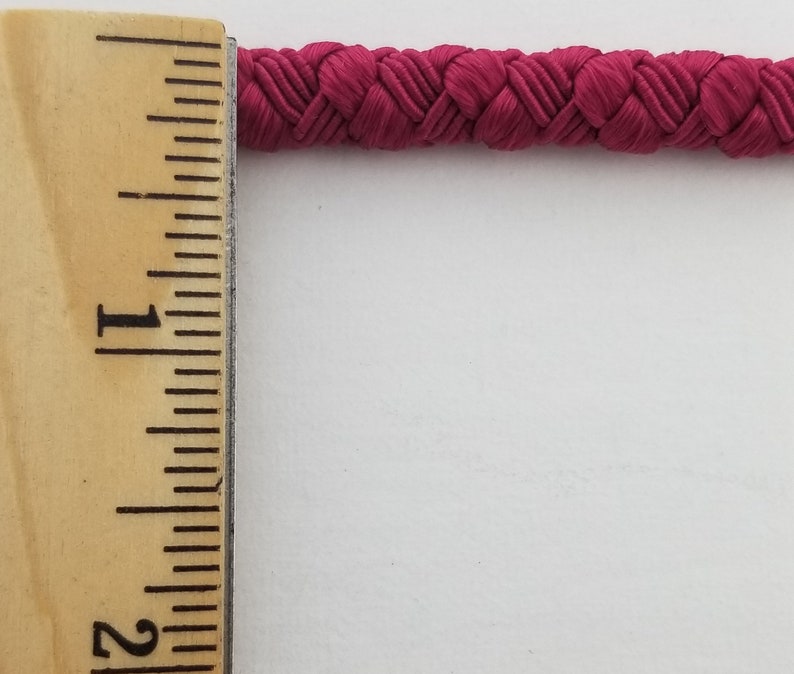 May include: A close-up of a 1-inch wide, crimson red braided cord. The cord is measured against a wooden ruler.