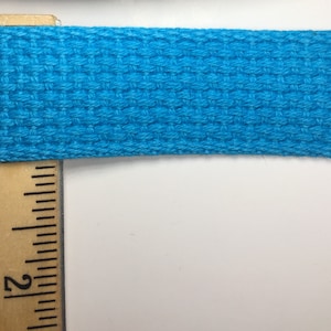 May include: A close-up of a bright turquoise woven cotton webbing, measured against a wooden ruler with the number "2" visible.