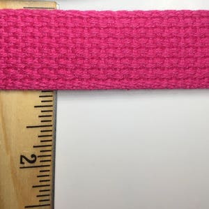 May include: A close-up of a pink woven ribbon or webbing, measured with a wooden ruler. The ruler shows the measurement of 2 inches.