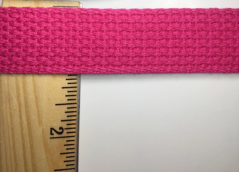 May include: A close-up of a pink woven ribbon or webbing, measured with a wooden ruler. The ruler shows the measurement of 2 inches.