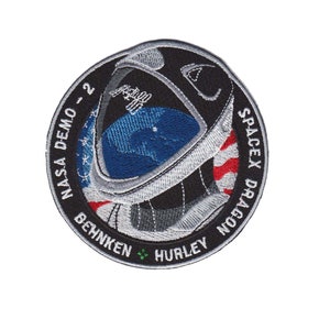 May include: A black and white embroidered patch with a silver astronaut helmet in the center. The patch is circular and has the text "NASA DEMO - 2" on the left side and "SPACEX DRAGON" on the right side. The text "BEHNKEN" and "HURLEY" are at the bottom of the patch.