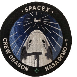 May include: A black and white embroidered patch with a white spacecraft against a blue background. The patch says "SpaceX" at the top, "Crew Dragon" on the left, and "NASA Demo-1" on the right.