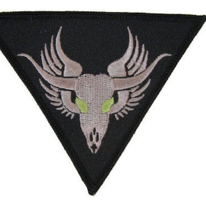 May include: A black triangular patch with an embroidered design of a grey skull with green eyes and wings. The skull is centered in the triangle and has a detailed design.