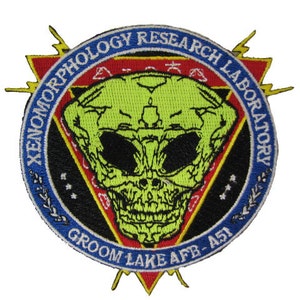 May include: A round, blue, white, and red embroidered patch with a green alien skull in the center. The text around the alien skull reads "XENOMORPHOLOGY RESEARCH LABORATORY GROOM LAKE AFB - 451".