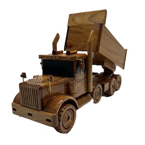 May include: A wooden toy dump truck with a raised bed. The truck is made of a light brown wood and has detailed features, including a front grill, headlights, and exhaust pipes. The bed is tilted upwards.