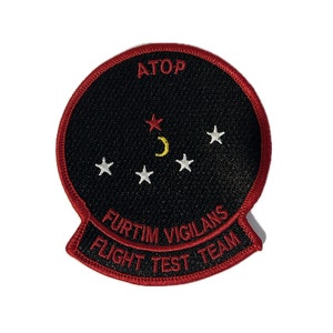 May include: A black and red embroidered patch with a white star and crescent moon design. The text "ATOP FURTIM VIGILANS FLIGHT TEST TEAM" is embroidered in red around the edge.