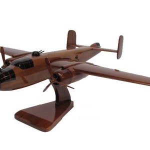May include: A wooden model of a brown B-25 Mitchell bomber airplane with a dark brown and black camouflage pattern. The model is displayed on a wooden stand.