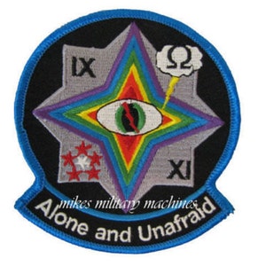 May include: A blue and black embroidered patch with a rainbow star, an eye, and the text "Alone and Unafraid". The star has the numbers IX and XI on it. The eye is surrounded by a rainbow and has a red slash through it. There is a lightning bolt and a cloud with the letter omega in it above the star.