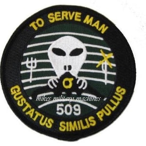 May include: A black and green embroidered patch with a white alien face in the center. The text "To Serve Man" is written in a yellow arc above the alien. The text "Gustatus Similis Pullus 509" is written in a yellow arc below the alien.