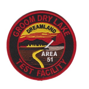 May include: A red, black, and yellow embroidered patch with the text "Groom Dry Lake Dreamland Area 51 Test Facility". The patch features a stylized image of a desert landscape with a road leading to a mountain range.