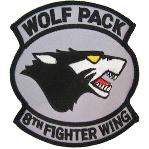 May include: A circular embroidered patch with a light gray background and a black border. The design features a black wolf head with a yellow eye and red mouth. The text WOLF PACK is arched at the top and 8TH FIGHTER WING is at the bottom.