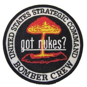 May include: A black and white circular patch with the text "United States Strategic Command Bomber Crew" around the edge. In the center is a red and yellow mushroom cloud with the text "got nukes?" underneath.