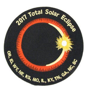 May include: A black and orange circular patch with the text "2017 Total Solar Eclipse" and a stylized image of the sun and moon. The text "OR, ID, WY, NE, KS, MO, IL, KY, TN, GA, NC, SC" is printed around the bottom of the patch.
