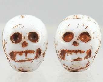 May include: Two white skull beads with brown painted details. The skulls have brown eyes and teeth.