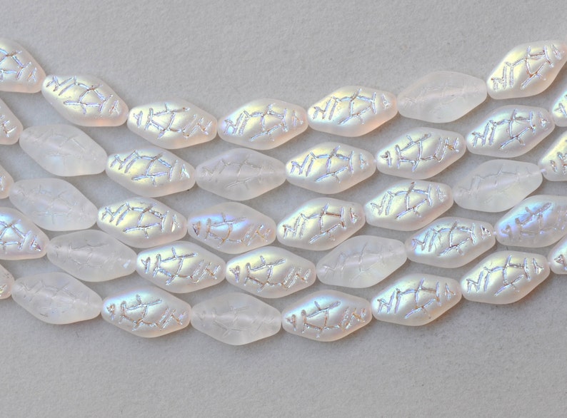 May include: Close-up of iridescent white oval beads with a textured, silver-colored design. The beads are arranged in rows, showcasing their unique shape and shimmering surface. Ideal for jewelry making.
