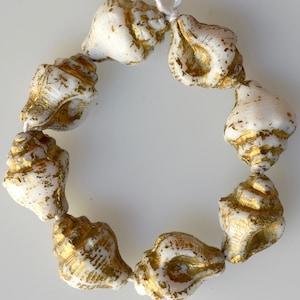 May include: A strand of white seashell beads with gold accents. The beads are strung together, forming a circular shape. The gold detailing adds a touch of elegance to the natural seashell design. Ideal for jewelry making or coastal decor.