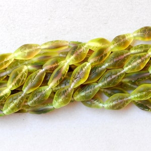 May include: Strands of olive green glass beads, each shaped like a leaf with etched vein details. The beads have a translucent quality, with hints of yellow and pink, strung together for jewelry making or crafts.
