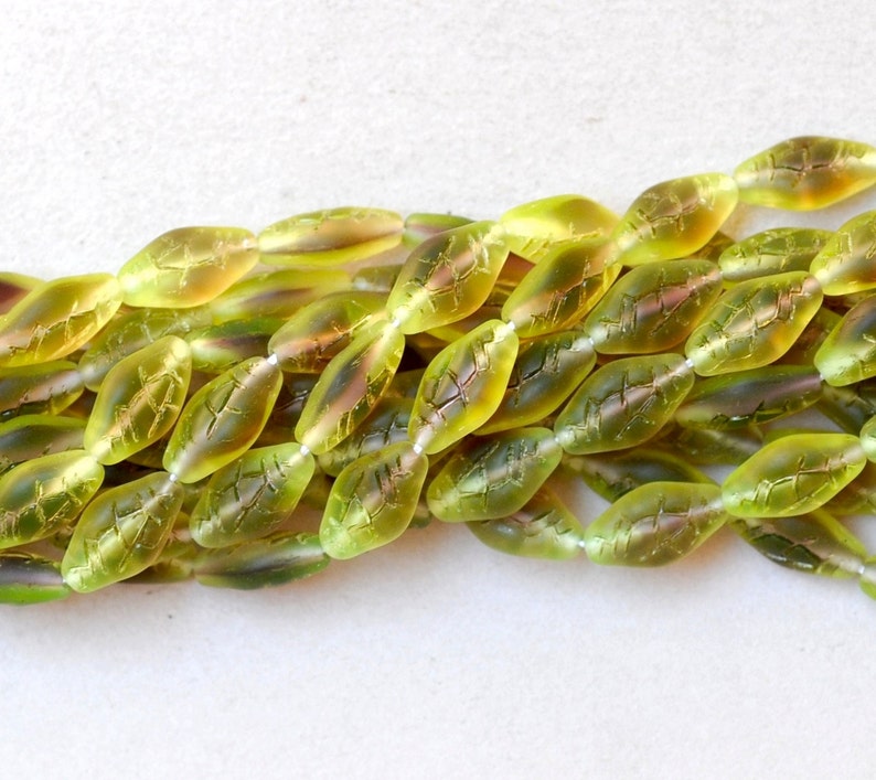 May include: Strands of olive green glass beads, each shaped like a leaf with etched vein details. The beads have a translucent quality, with hints of yellow and pink, strung together for jewelry making or crafts.