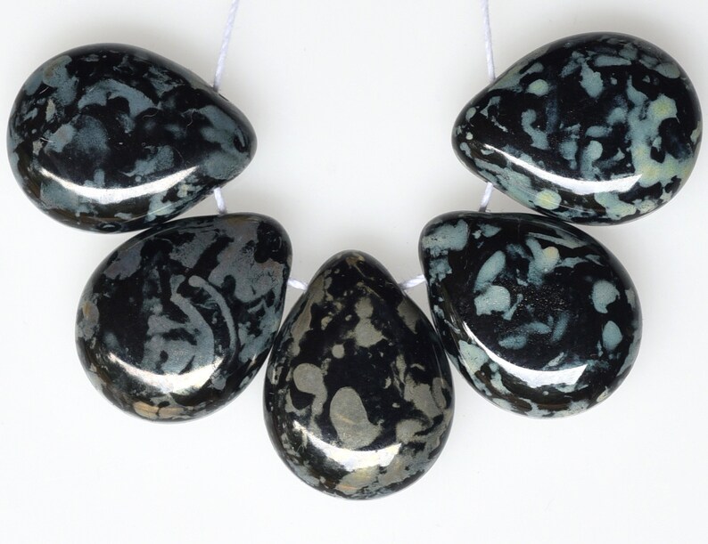 May include: A strand of five teardrop-shaped beads. The beads are primarily black with a pattern of gray and white inclusions. They are strung on a white cord, and the beads have a polished, glossy finish.