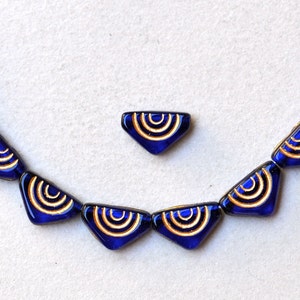 Puede incluir: Un collar hecho de cuentas de vidrio azul con detalles dorados. Cada cuenta tiene forma de triángulo con bordes redondeados y presenta un diseño de círculos concéntricos. El collar está enhebrado en un hilo transparente y tiene un colgante a juego.