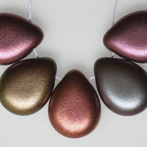 May include: A strand of six metallic teardrop-shaped beads in shades of bronze, gold, and pink. The beads are strung on a white cord, and are displayed against a white background. The beads have a smooth, iridescent finish.