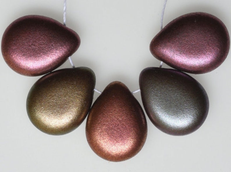 May include: A strand of six metallic teardrop-shaped beads in shades of bronze, gold, and pink. The beads are strung on a white cord, and are displayed against a white background. The beads have a smooth, iridescent finish.