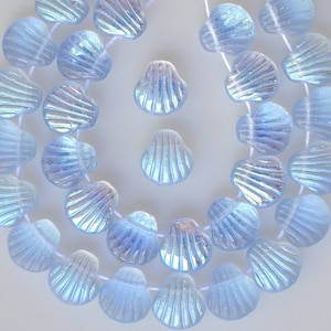 Puede incluir: Una hebra de cuentas de vidrio azul iridiscente en forma de concha. Las cuentas están enhebradas en un hilo blanco y están dispuestas en un patrón circular. Las cuentas son todas del mismo tamaño y forma, y tienen un acabado ligeramente iridiscente.