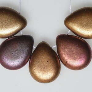 May include: Five metallic teardrop-shaped beads strung on a white cord. The beads have a textured surface and vary in color from gold to bronze and reddish-purple. These beads could be used for jewelry making.