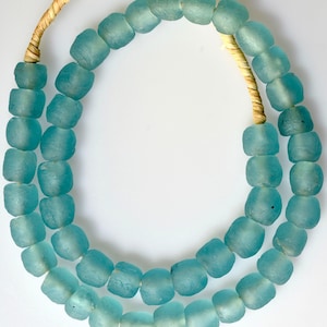 May include: A necklace featuring two strands of light blue, square-shaped beads. The beads have a frosted appearance and are strung on a natural-colored, woven cord. The necklace is displayed against a white background.