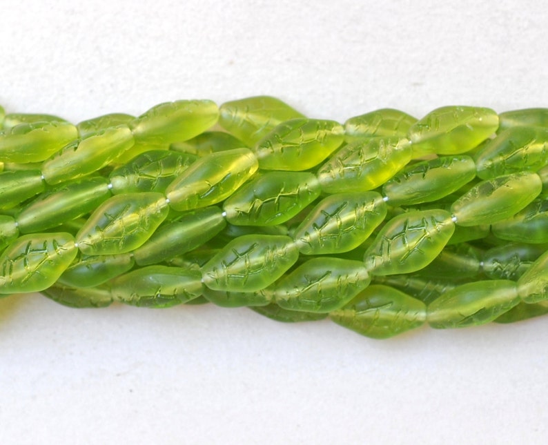 May include: Close-up of strands of translucent, lime green leaf-shaped beads. Each bead has etched leaf vein details. The beads are strung together, creating a textured and organic aesthetic. Ideal for jewelry making.